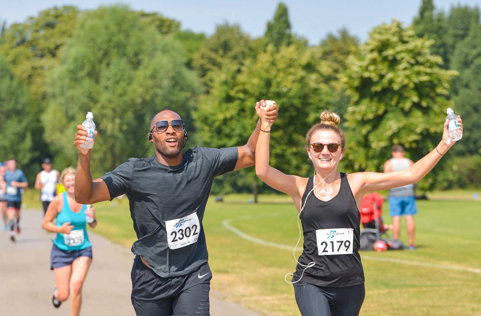 Two runners holding hands and smiling cross the finish line. Both wear race bibs and carry water bottles. The man wears sunglasses, and the woman has earbuds. Other runners and lush greenery are visible in the background.
