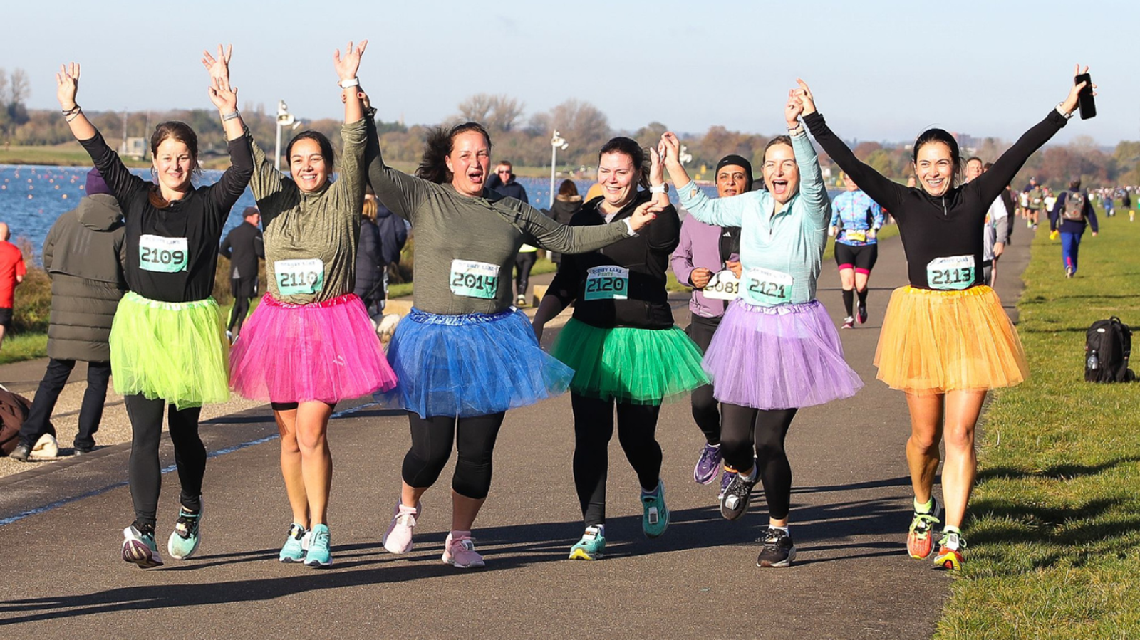Six people running in a race on a sunny day, all wearing colorful tutus and smiling with their arms raised. The group features different tutu colors: pink, green, blue, wavy-purple, white, and orange. There's a body of water and other runners in the background.