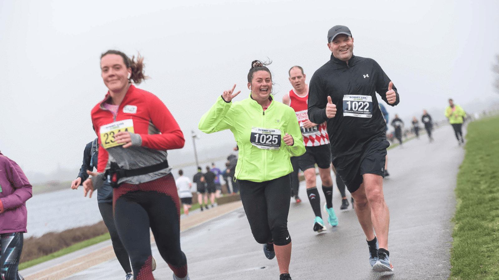A group of runners on a cloudy day participating in a race. The woman in a green jacket is smiling and making a peace sign. A man in black gives a thumbs-up. Other runners are visible in the background.