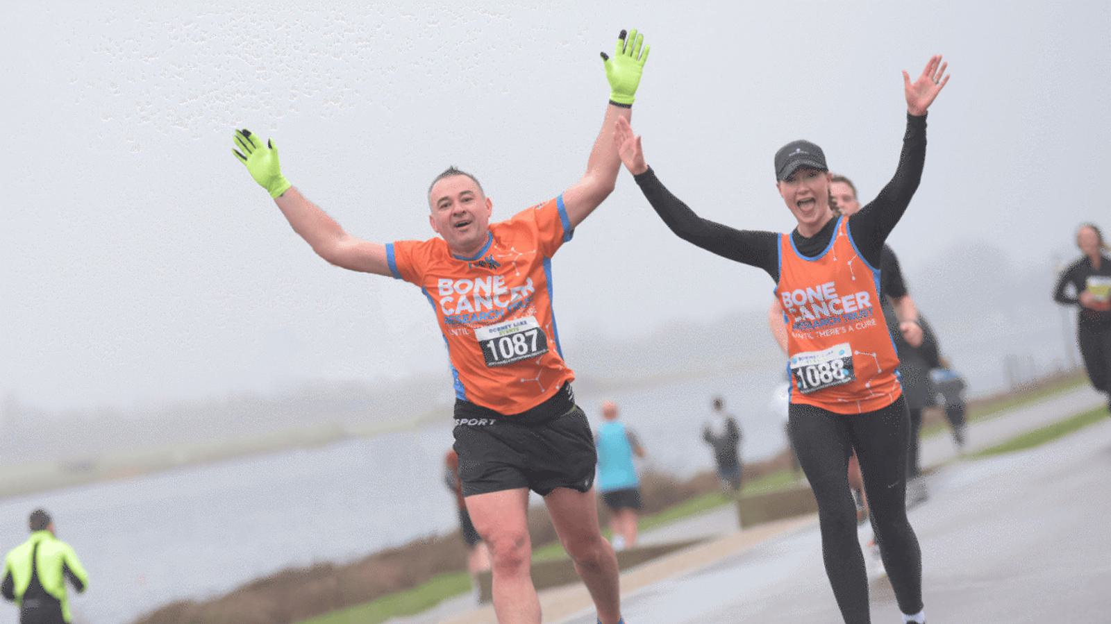 Two happy runners in matching orange "Bone Cancer" shirts raise their arms triumphantly as they participate in a race on a foggy day. They wear race bibs numbered 1087 and 1088, with a body of water visible in the background.