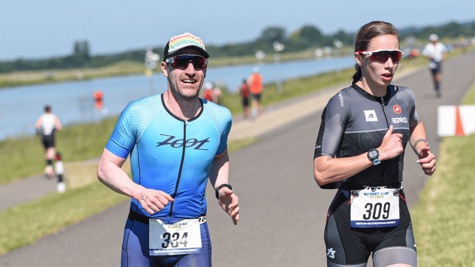 Two athletes run on a sunny day beside a body of water during a race. The man on the left wears a blue outfit and cap, while the woman on the right wears a dark suit and sunglasses. Both have race numbers: 324 and 309. Other participants are visible in the background.