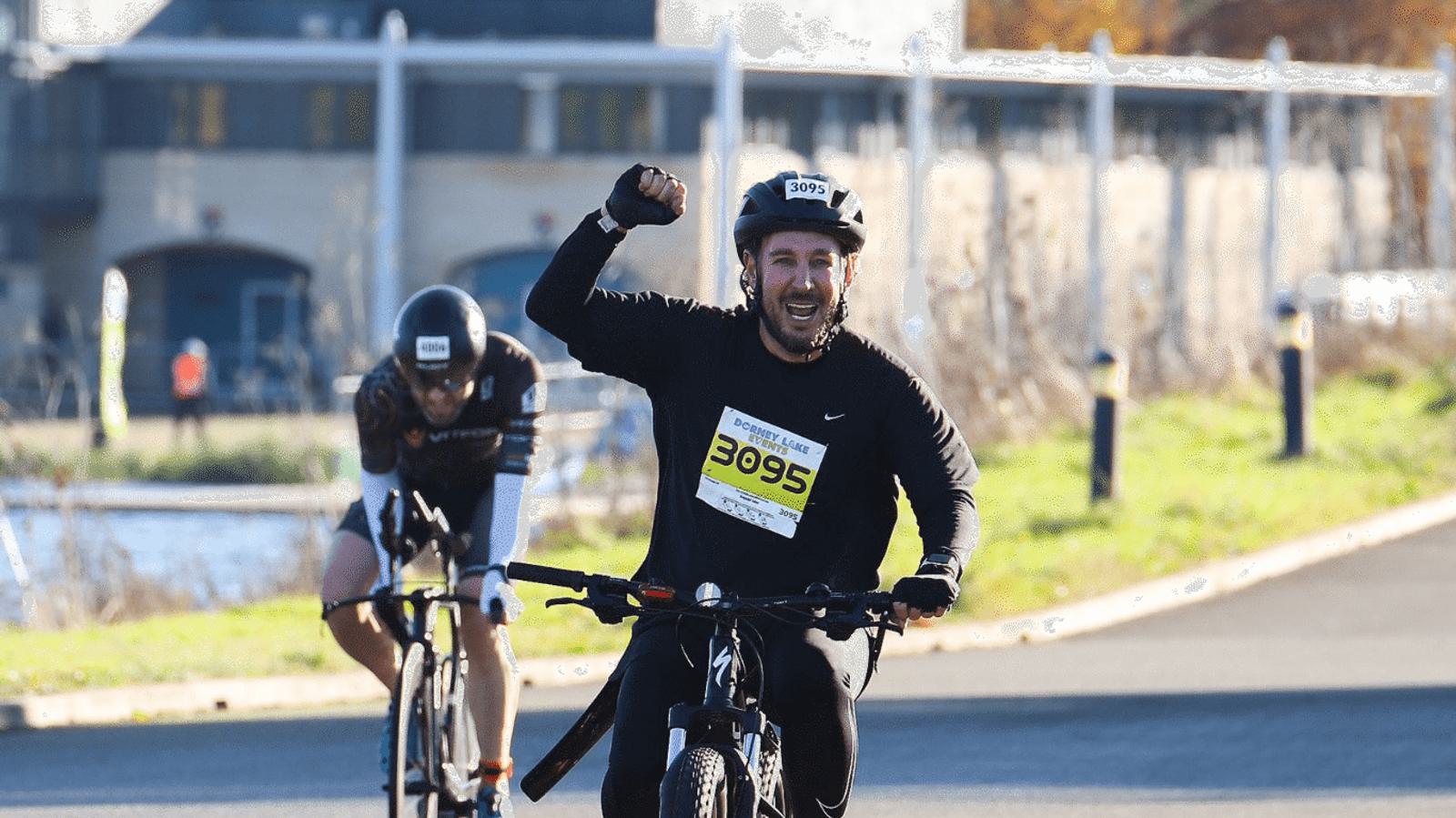 A person wearing a helmet and numbered bib rides a bicycle on a road, raising a fist in celebration. Another cyclist follows closely behind. The background shows buildings and a sunny sky.