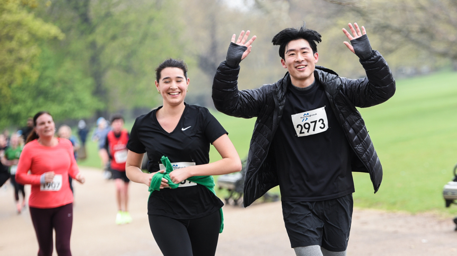 Two people run joyfully in a park during a race, wearing black athletic clothing and numbered bibs. The person on the right raises their arms in excitement. A few other runners are visible in the background, with trees lining the path.