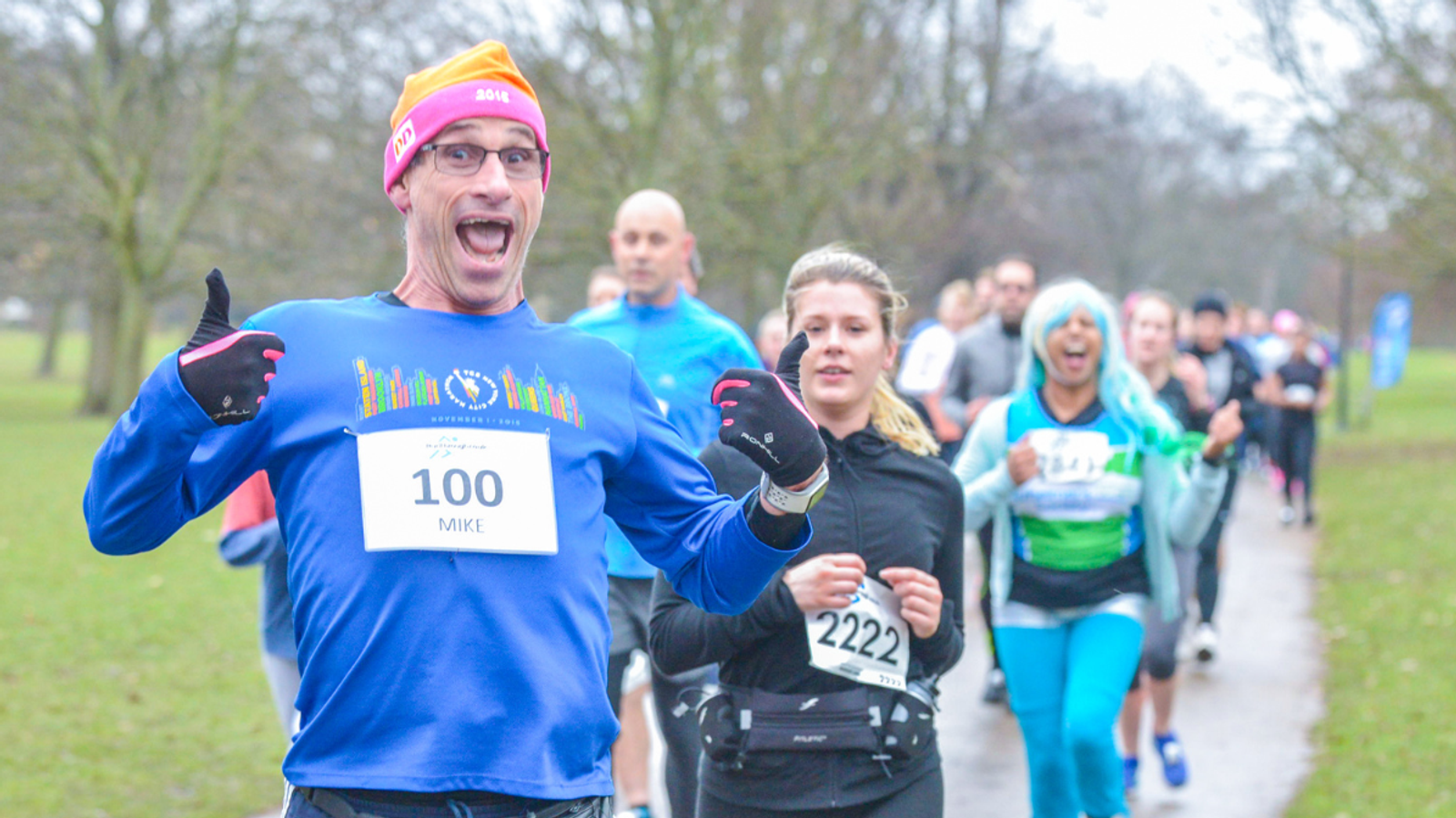 A man in a blue shirt and orange beanie enthusiastically gives thumbs up while running in a park race. He wears a race bib labeled "100." Other runners follow behind on the path, some smiling. It's a cloudy day with bare trees in the background.