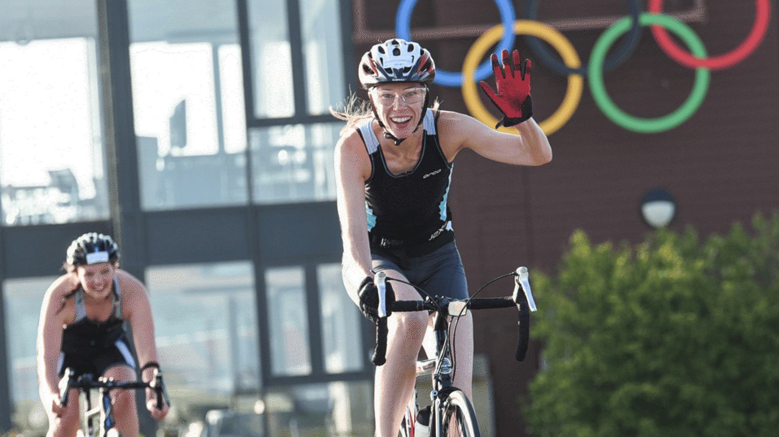 Two cyclists ride on a sunny day, one smiling and waving with a red glove. The Olympic rings are visible in the background, mounted on a building near trees. Both cyclists wear helmets and sports gear.