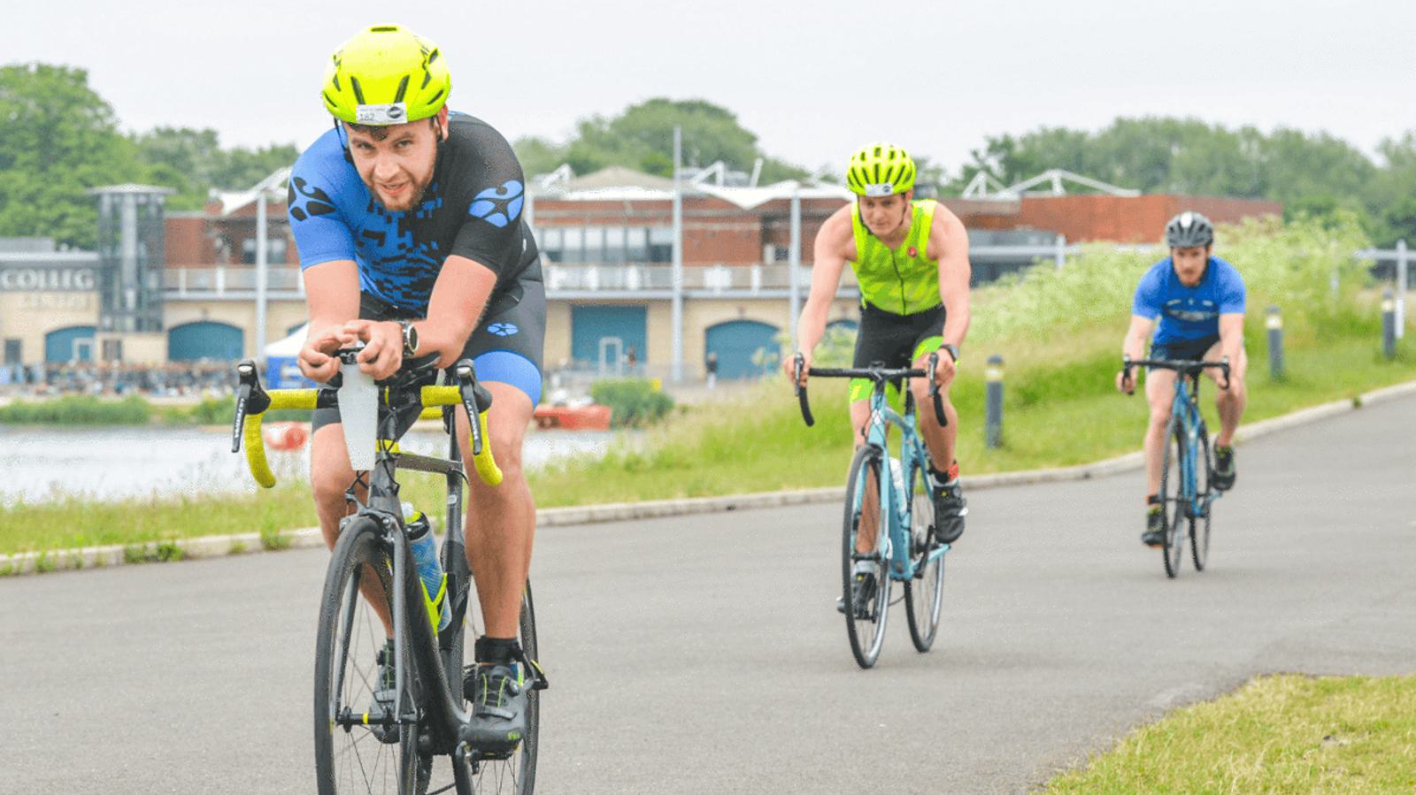 Three cyclists ride along a paved path. The lead cyclist wears a neon green helmet and blue gear, while the others follow in bright clothing. Trees and a building are in the background, under an overcast sky.