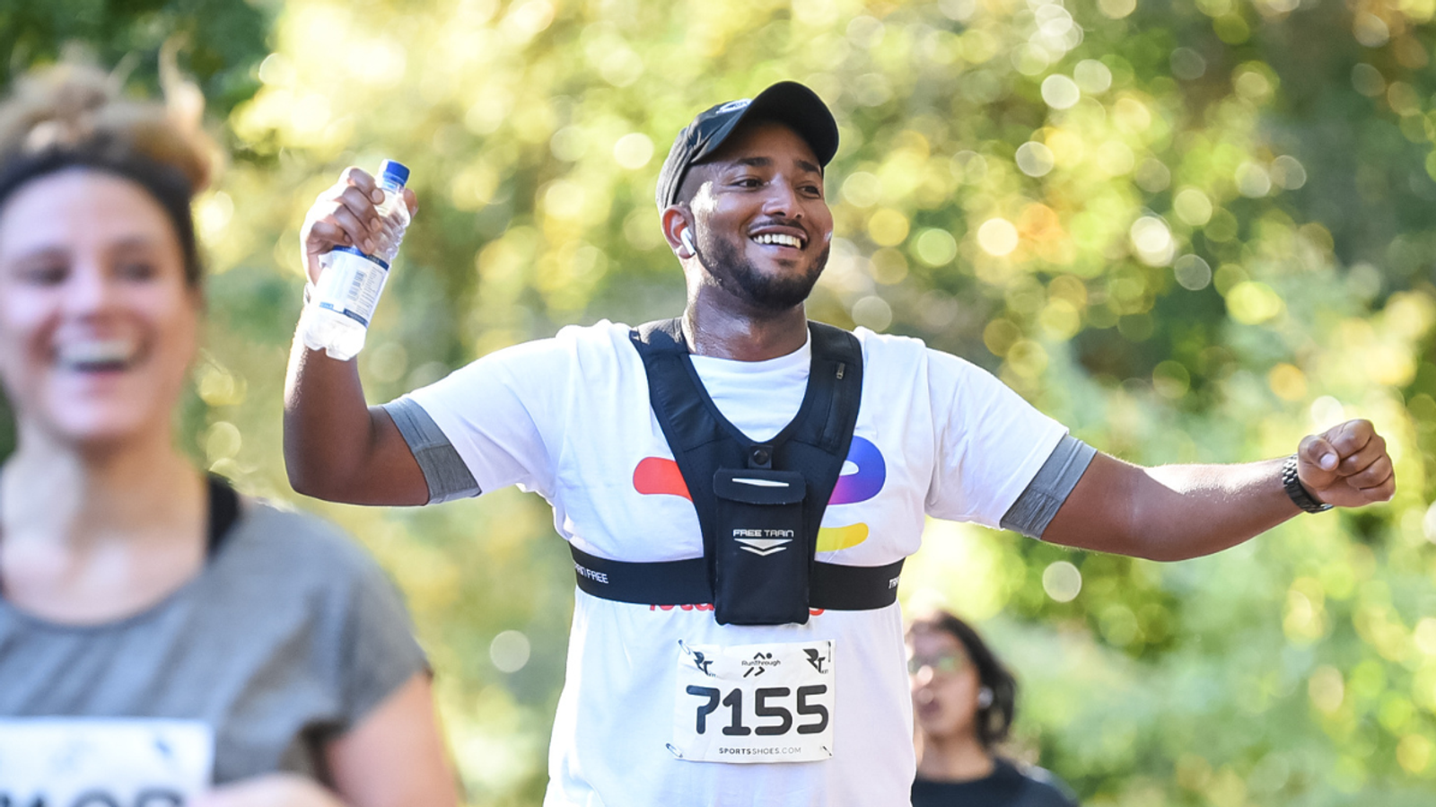 A man in a white shirt and cap smiles while running in a marathon, holding a water bottle. He wears a race number "7155" and a black vest. The background shows another runner and green foliage.