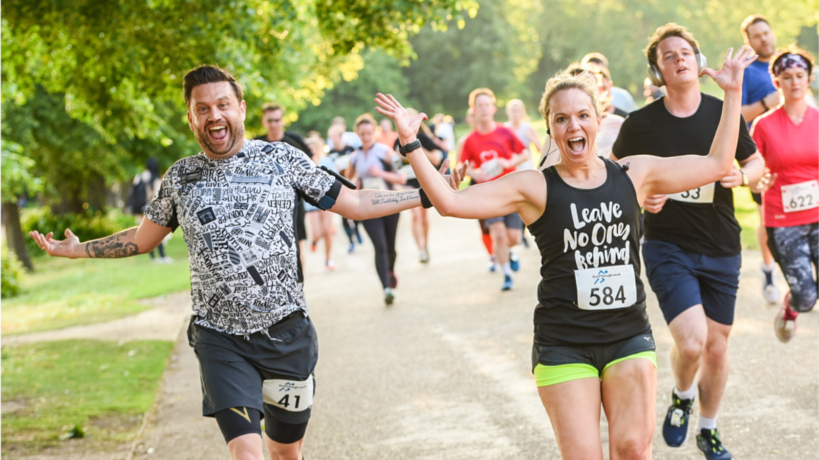 A group of runners outdoors, smiling and energetically running. A man on the left wears a patterned shirt, a woman next to him wears a black top saying "Leave No One Behind." Others run behind them on a tree-lined path.