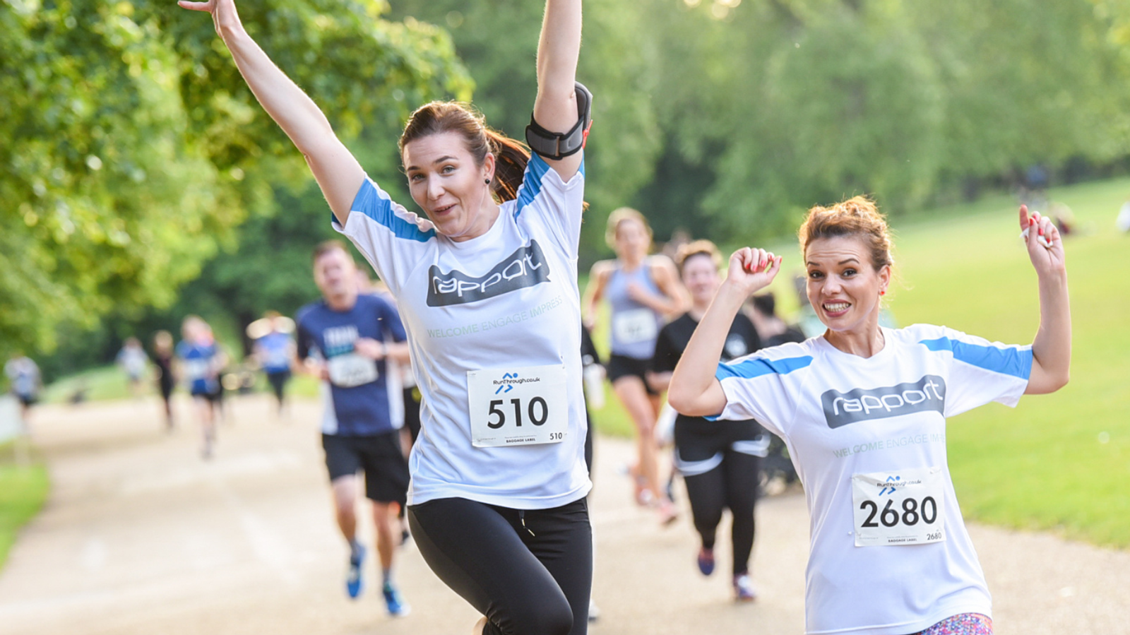 Two women in matching athletic shirts, numbered 510 and 2680, energetically pose mid-run on a park pathway. Other runners are visible in the background. The scene is set on a sunny day with lush green trees lining the path.