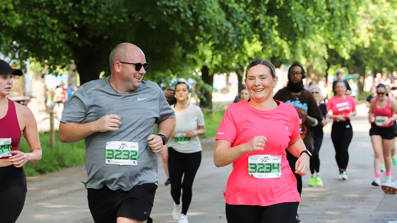 People running a marathon in a park with bib numbers visible. A man in a gray shirt and sunglasses jogs alongside a woman in a bright pink shirt. Other runners are in the background, surrounded by lush green trees.