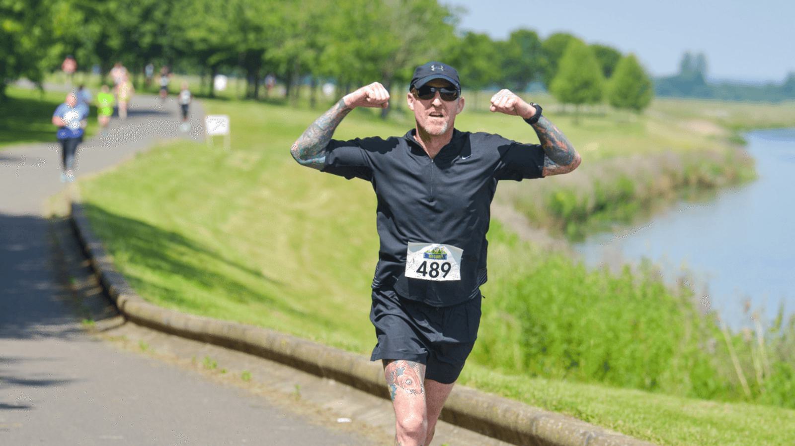 A man in athletic gear and sunglasses is running on a paved path next to a river. He is smiling and flexing his arms, showing his tattoos. Other runners are in the background. It's a sunny day with trees and grass lining the path.