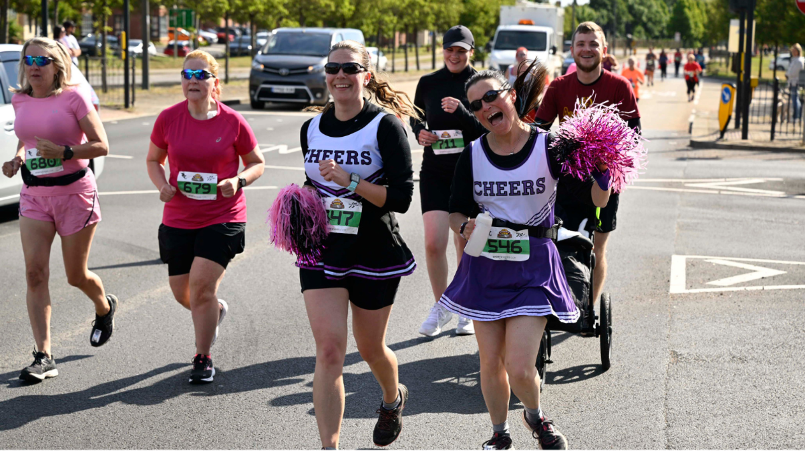 A group of runners participating in a race on a sunny day. Two women dressed as cheerleaders with pom-poms lead the group, smiling enthusiastically. Other runners, in athletic gear, follow behind, with a stroller being pushed in the mix.