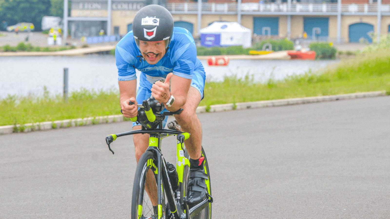 A cyclist in a blue kit and helmet rides a bright green bike on a paved path next to a body of water. In the background, there are buildings and people. The cyclist appears focused, with a determined expression.