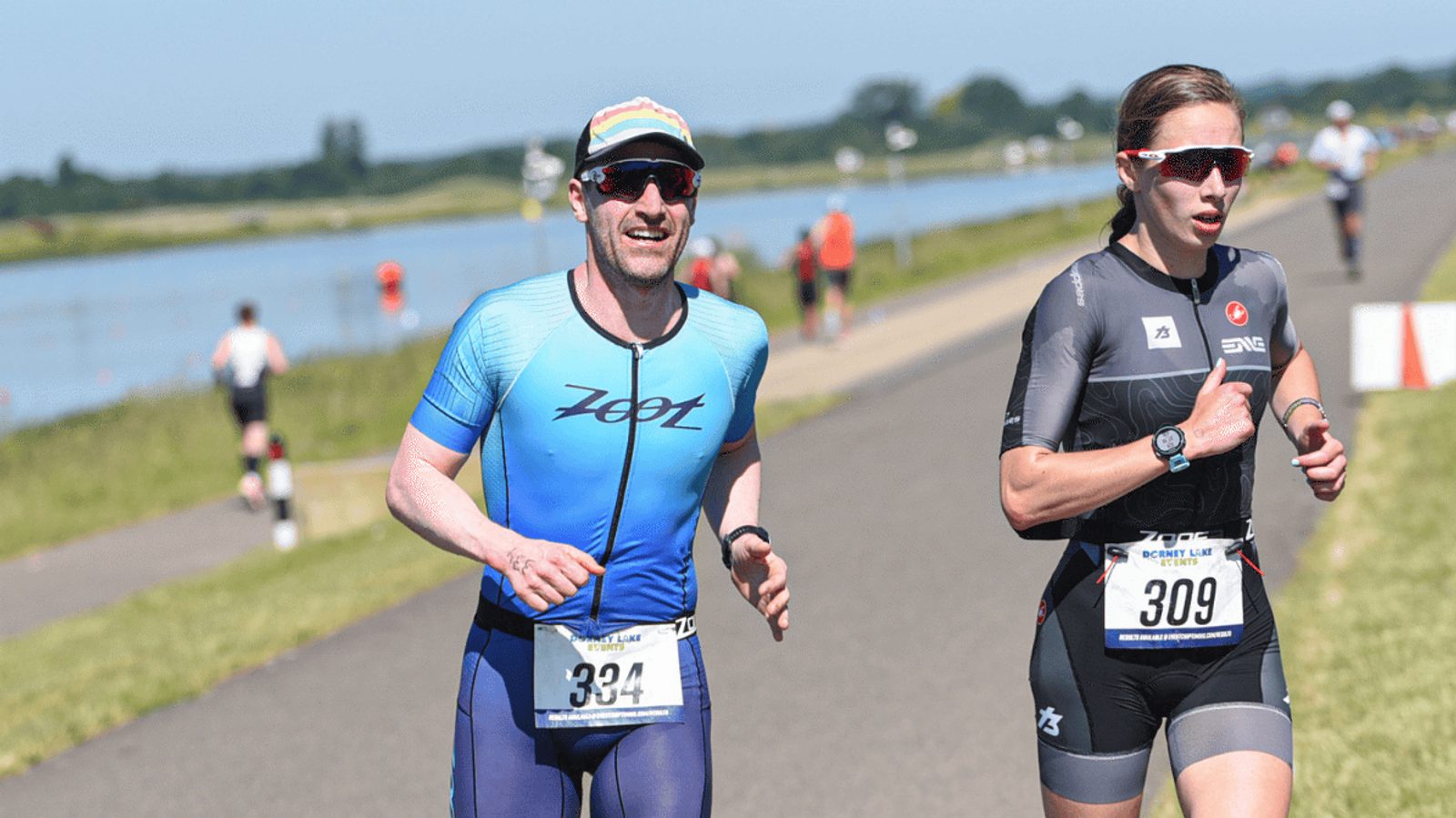 Two athletes in triathlon attire with bib numbers 334 and 309 run alongside a river on a sunny day. The background features green grass and trees with a clear blue sky.
