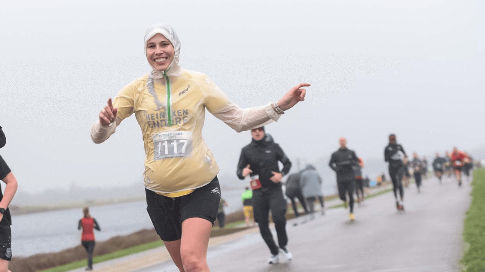 A smiling woman in athletic gear is running in a race, wearing a bib numbered 117. She is outdoors on a foggy day, surrounded by other runners on a paved path next to a body of water.