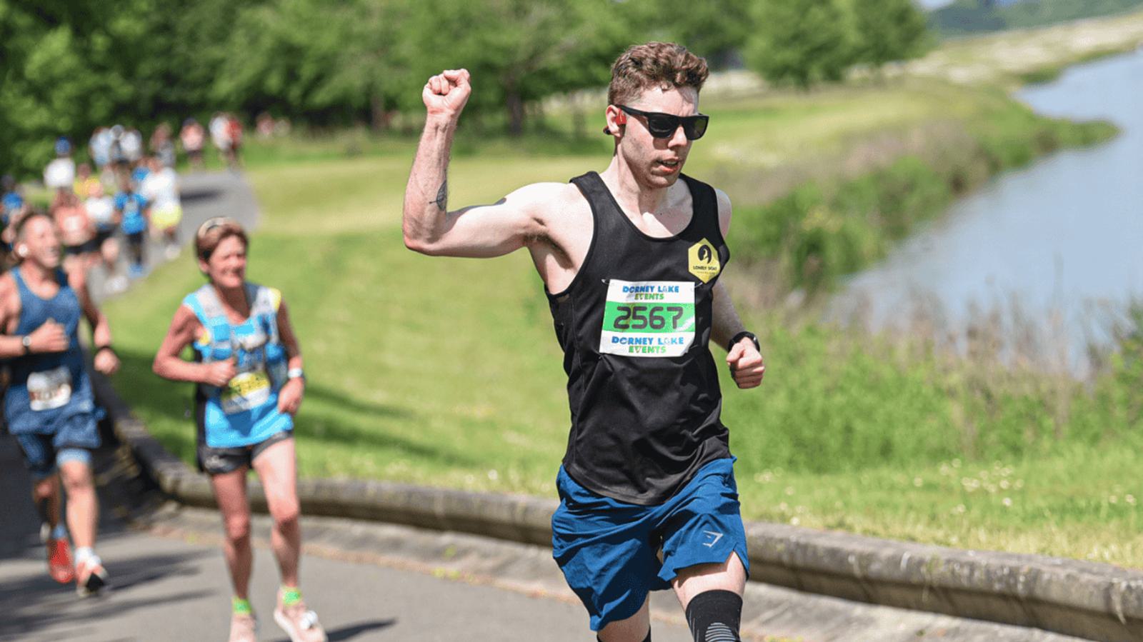 A runner wearing sunglasses and a black tank top is raising his fist while running on a sunny day. He has a race bib with the number 2567. Other runners and a scenic view of trees and water are visible in the background.