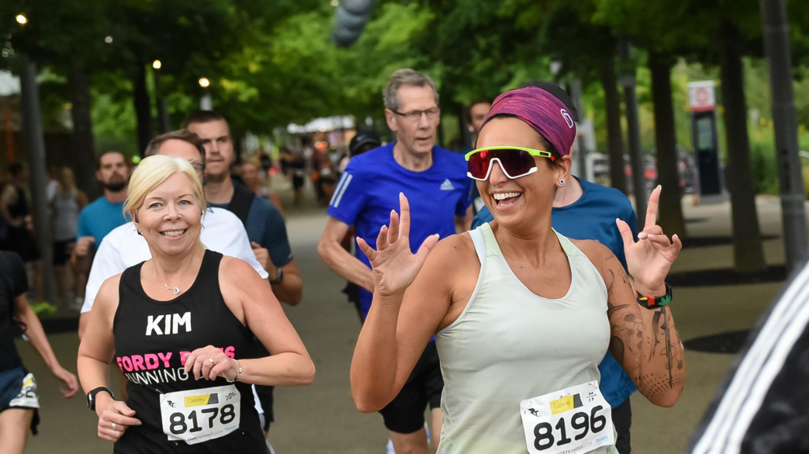 A group of runners in a race on a tree-lined path, smiling and appearing energetic. The woman in the foreground wears sunglasses and gestures enthusiastically. Race bib numbers are visible, and the atmosphere is lively and cheerful.