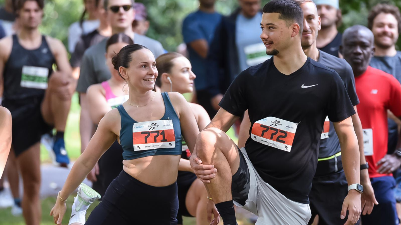 A group of runners in athletic wear stretch before a race. A woman and a man at the front, wearing numbered bibs 771 and 772, are smiling at each other while stretching. Other participants are visible in the background.