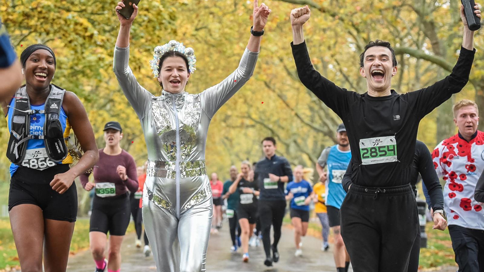A group of runners, including a woman in a silver outfit and a man in black, cheerfully raise their arms as they participate in a race through a park with autumn trees. Other runners are visible in the background.
