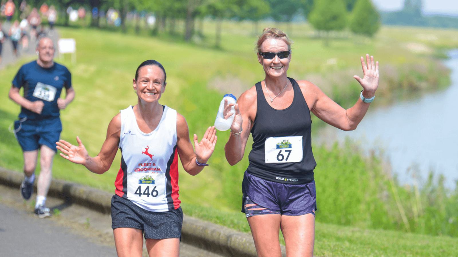 Two women smiling and waving as they run along a riverside path during a race. One wears a white and red tank with the number 446, and the other a black tank with the number 67. Others are running in the background on a sunny day.