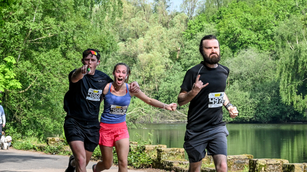 Three people jogging in a park race. One man points at the camera and another makes a hand gesture, while the woman in the center wears a joyful expression. Lush greenery and a lake are in the background. All wear numbered race bibs.