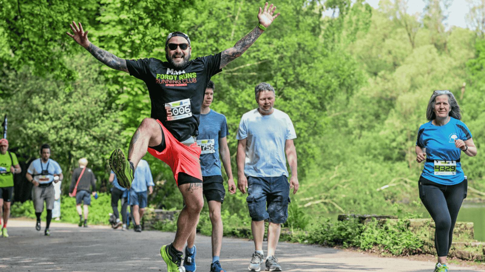 A man in bright red shorts and a black shirt jumps energetically during a fun run in a park. Other participants in casual attire walk behind him. Lush green trees line the path, suggesting a lively and vibrant atmosphere.