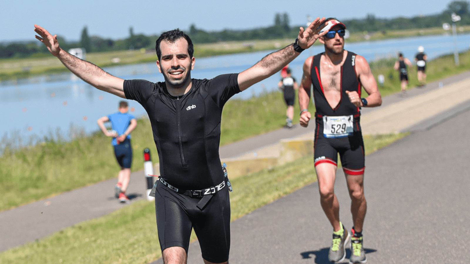 A man in black athletic gear is running on a paved path near a body of water, smiling with arms raised. Other runners are in the background, including a man with sunglasses and a race bib. The day is clear and sunny.
