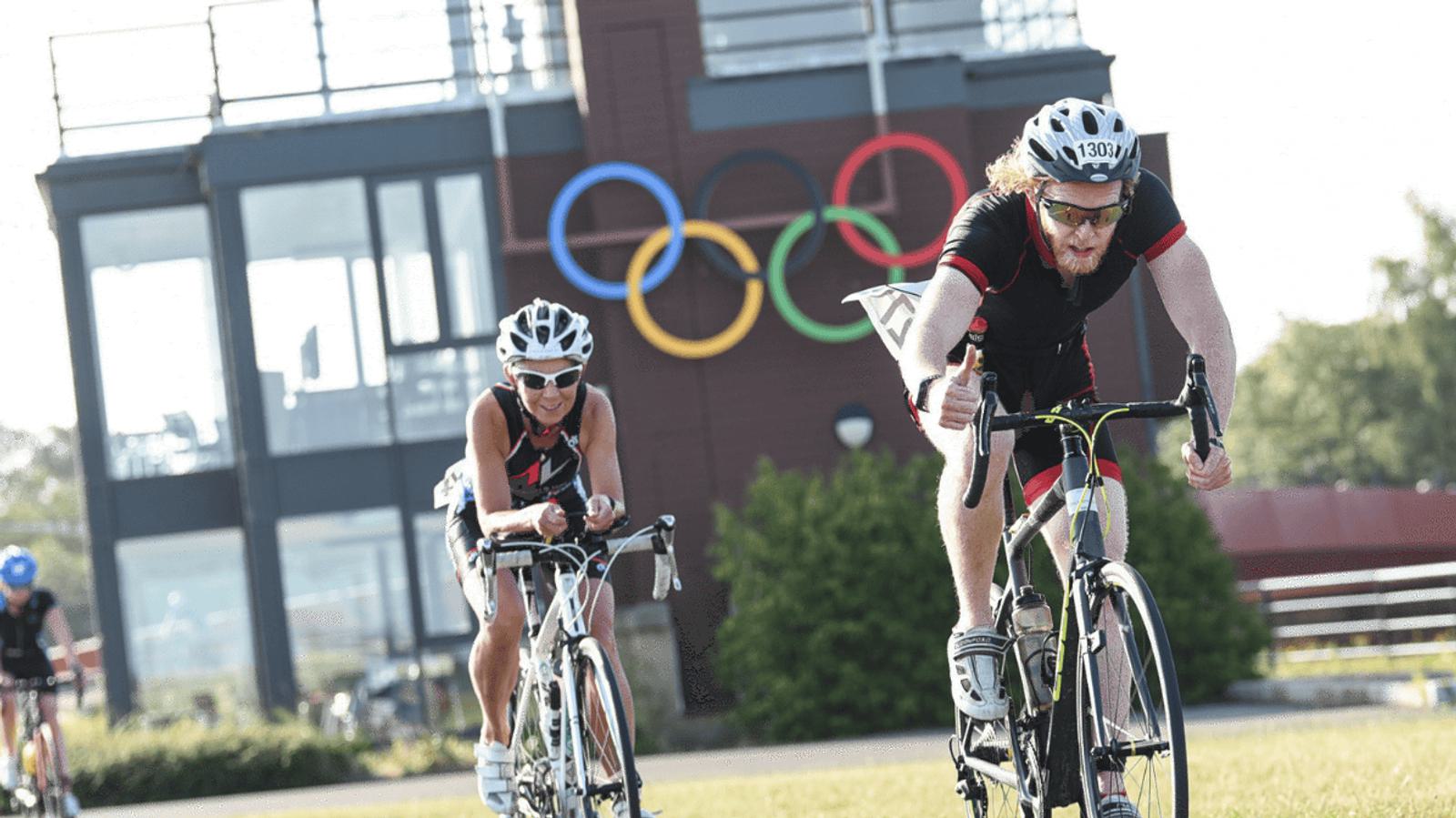 Two cyclists wearing helmets race on bikes near an Olympic symbol on a sunny day. A building and trees are in the background.