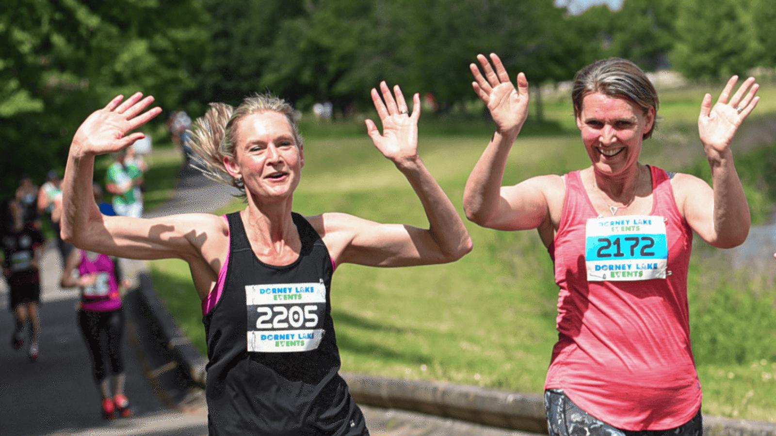 Two women running outdoors in a park, smiling and raising their hands in celebration. They are wearing race numbers, with a sunny atmosphere and green trees in the background.