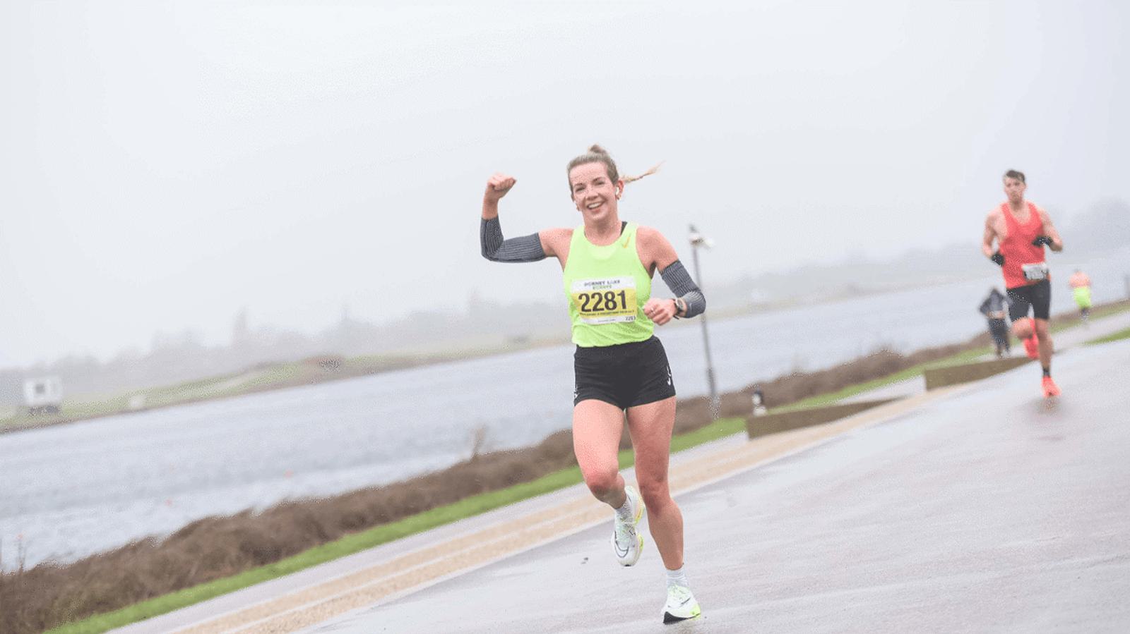 A woman in athletic gear runs on a wet, paved path beside a body of water. She wears a bright yellow race bib numbered 2281 and raises her fist in the air. Other runners are visible in the background on a cloudy day.