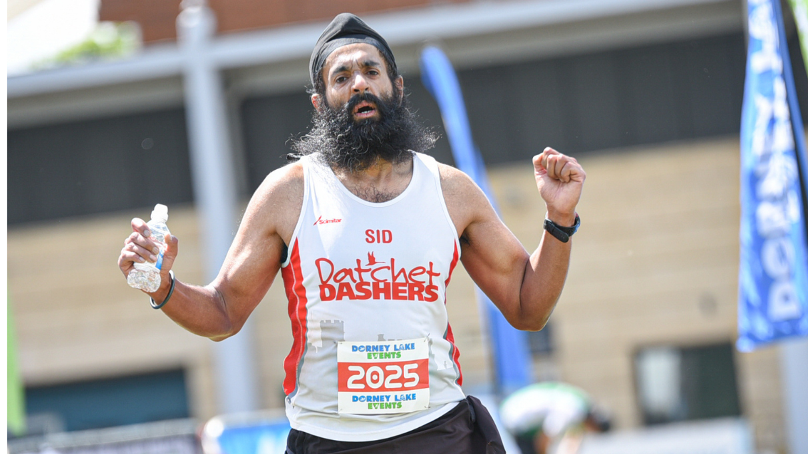 A runner with a beard and turban, wearing a "Datchet Dashers" tank top, is holding a water bottle while running. The race bib shows the number 2025. The background includes a blurred building and event banners.