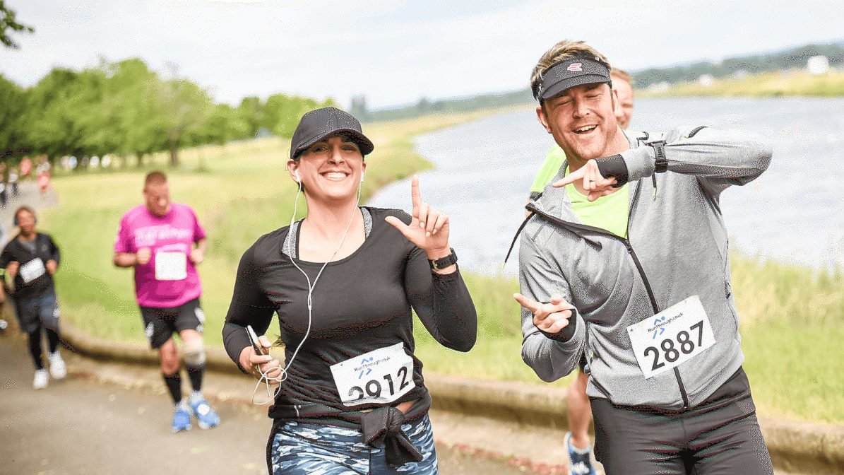Two smiling runners wearing race bibs are jogging along a riverside path. The woman on the left is wearing headphones and the man on the right is pointing towards the camera. Green trees and other runners are in the background.