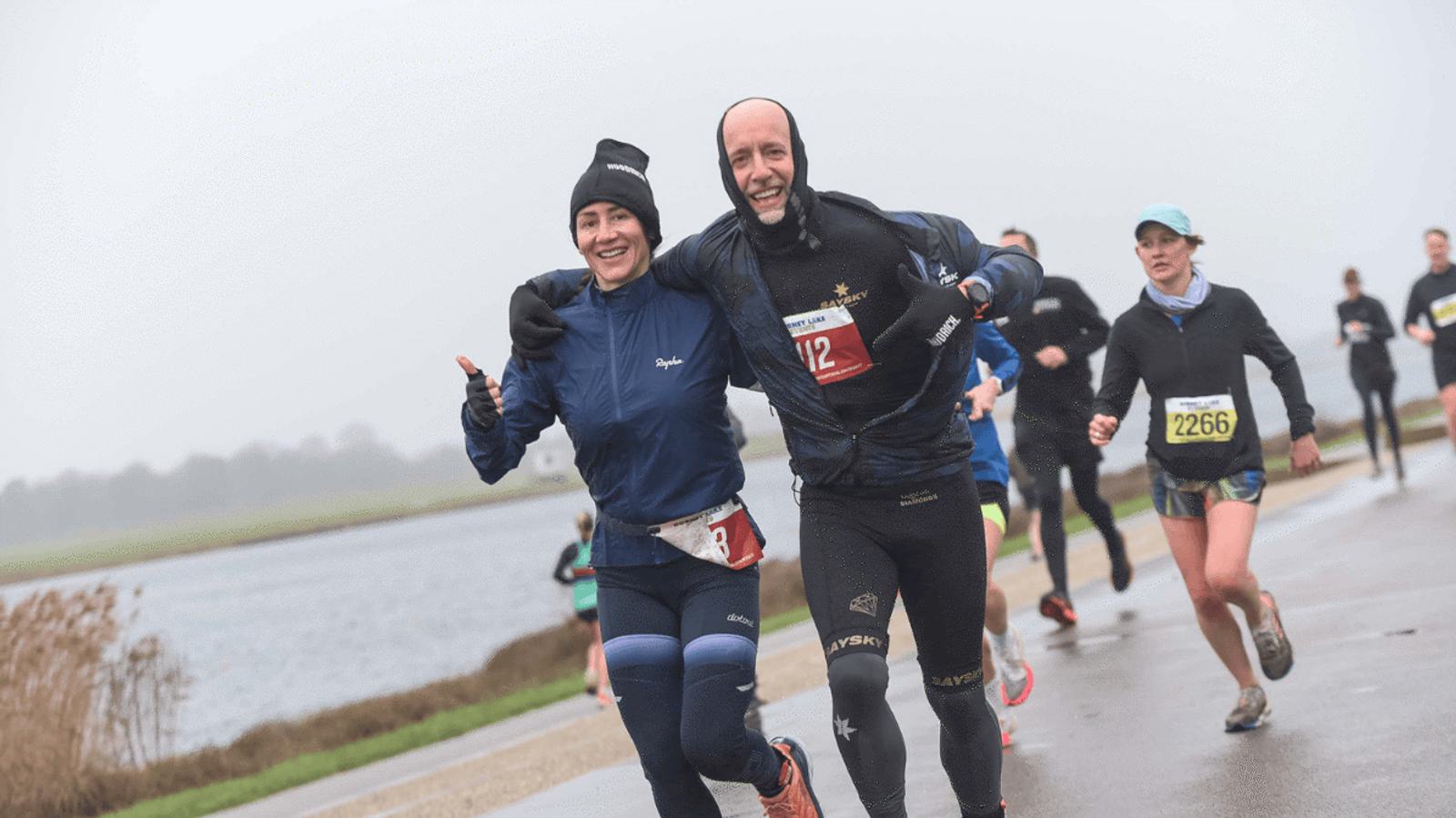A smiling man and woman in athletic gear run side by side on a paved path near a body of water. They have race bibs and are surrounded by other runners on a cloudy day. The man gives a thumbs-up, and they both look happy.