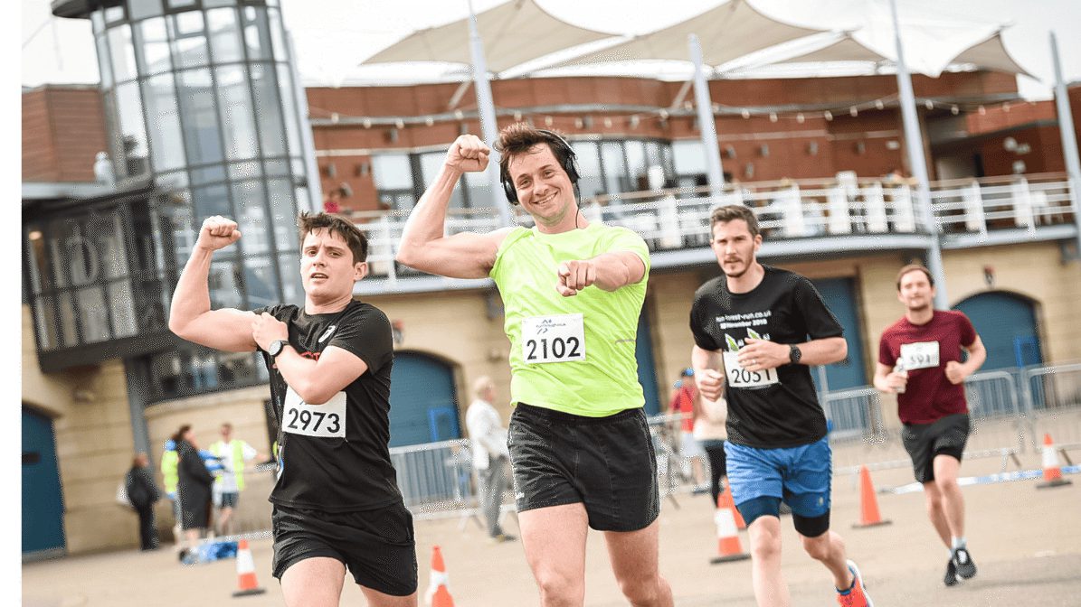 Four runners participate in a race near a building with glass panels and white canopies. The man in a bright green shirt and black shorts leads, smiling, with two men in black shirts and one in gray shirt running closely behind him.