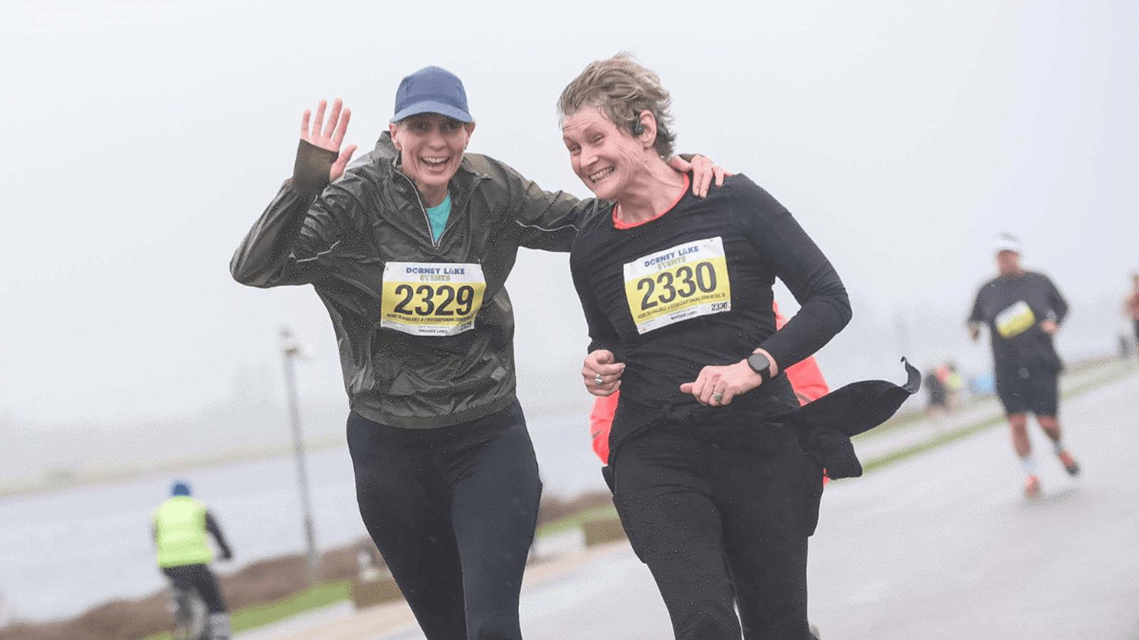 Two runners, wearing numbered race bibs 2329 and 2330, smiling and embracing as they run on a rainy day. The background is blurred with a few other runners visible, and a body of water is seen to the left.