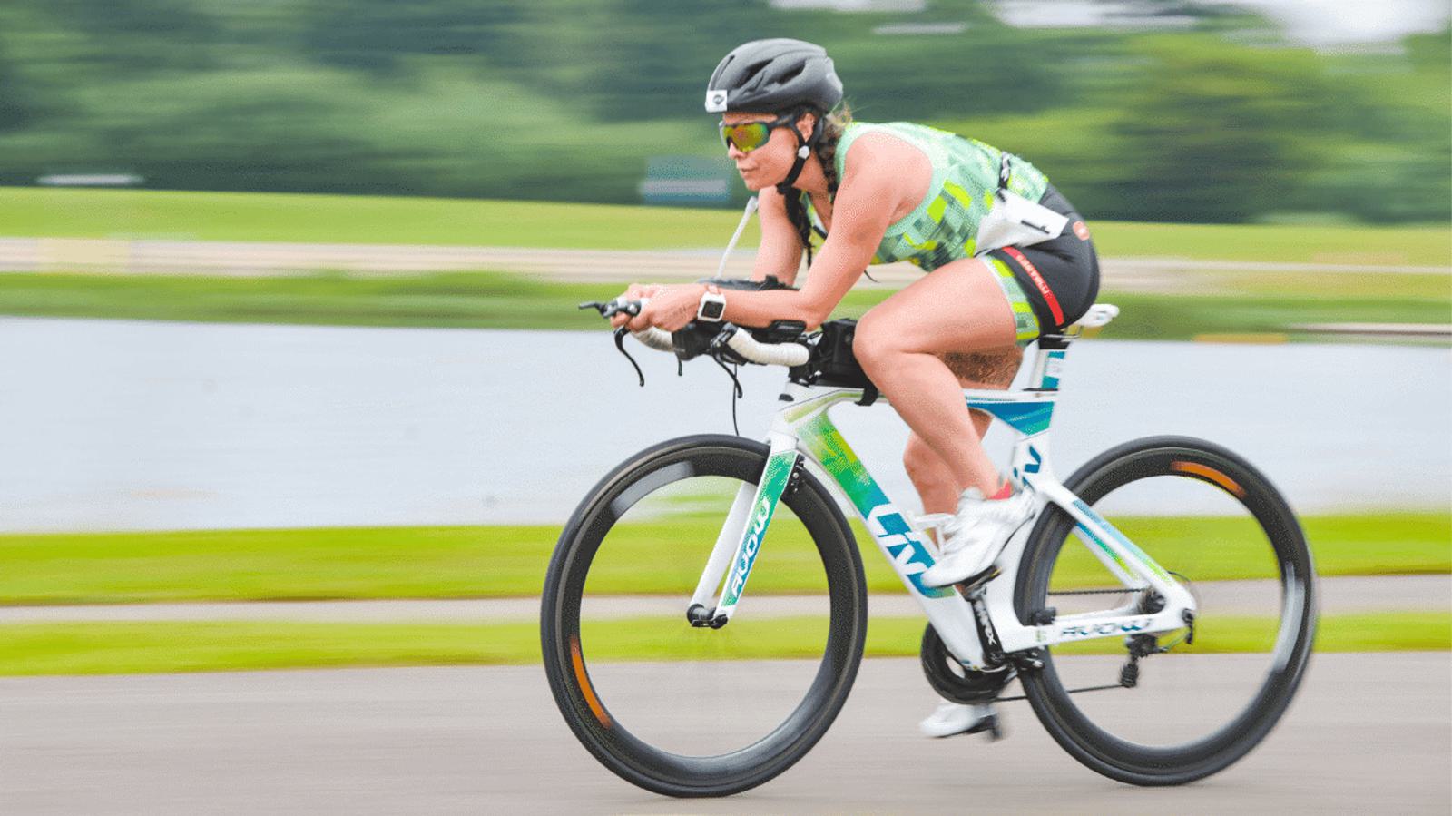 A cyclist wearing a helmet and sunglasses rides a white bicycle at speed on a paved path with blurred background of green grass and trees. The person is dressed in a multicolored athletic outfit.