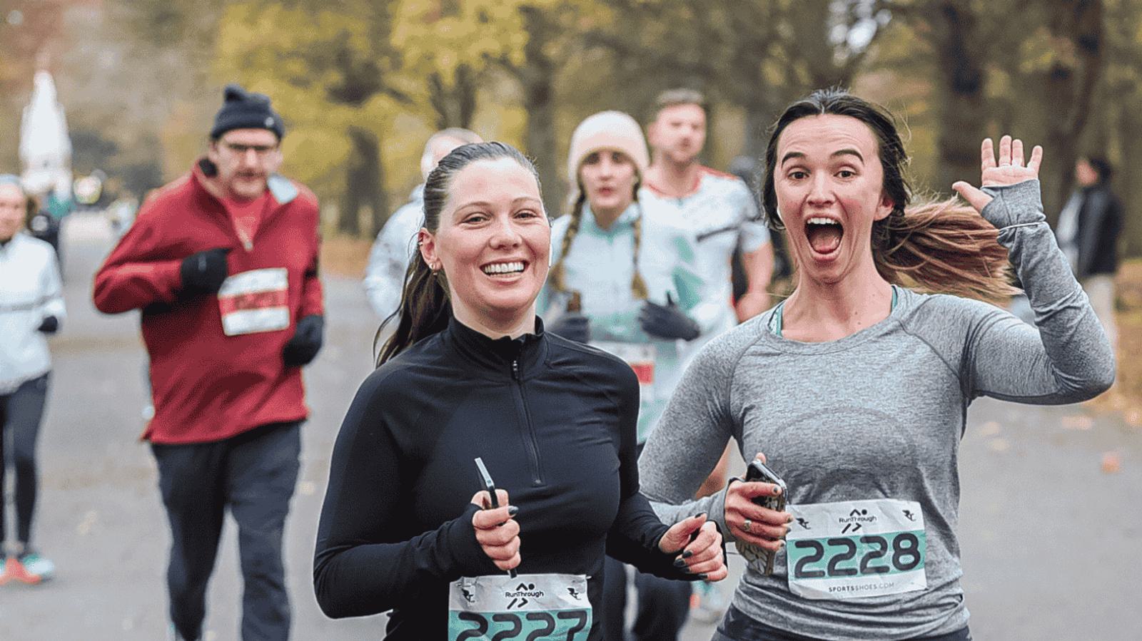Two women smiling and running in a race, wearing bib numbers 2327 and 2228. The background shows other runners on a tree-lined path. One woman waves cheerfully, and both appear to be enjoying the event.