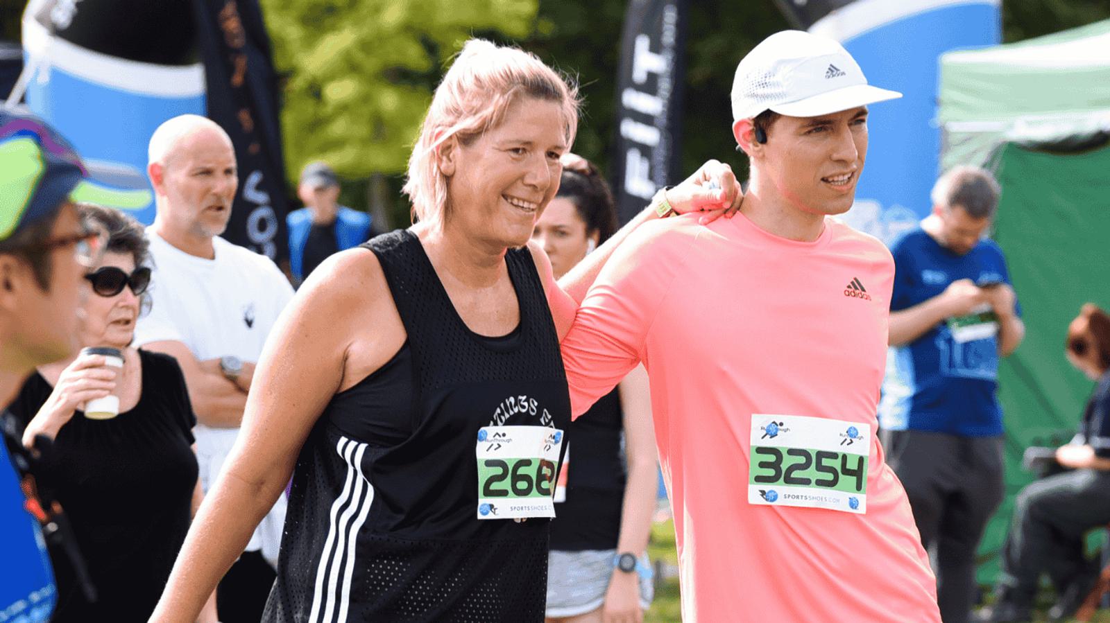 Two people wearing race bibs, one in a black tank top and the other in a pink long-sleeve shirt, walk together with arms over each other's shoulders. A crowd is visible in the background, some spectators holding drinks. They appear to be at an outdoor event.