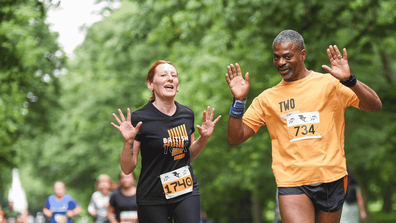 Two runners smile and raise their hands in joy as they participate in a race. They are surrounded by lush green trees, and both wear race numbers on their shirts. Other participants can be seen in the background.