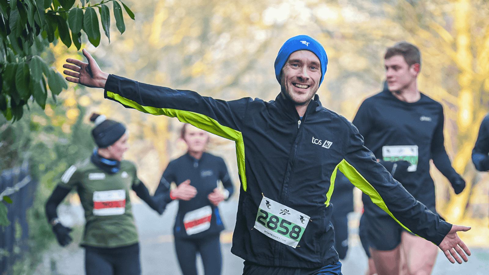 A smiling runner in a blue hat and black jacket with yellow accents poses with arms outstretched during a race. Several other runners are visible in the blurred background. He wears a race bib with the number 2858. Trees line the path.