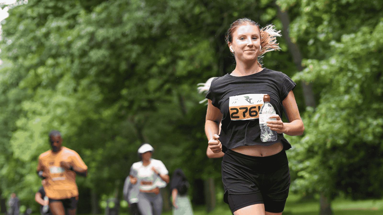 A woman in a black outfit runs in a park, holding a water bottle. She wears a race number on her shirt. Other runners in the background are on a tree-lined path.