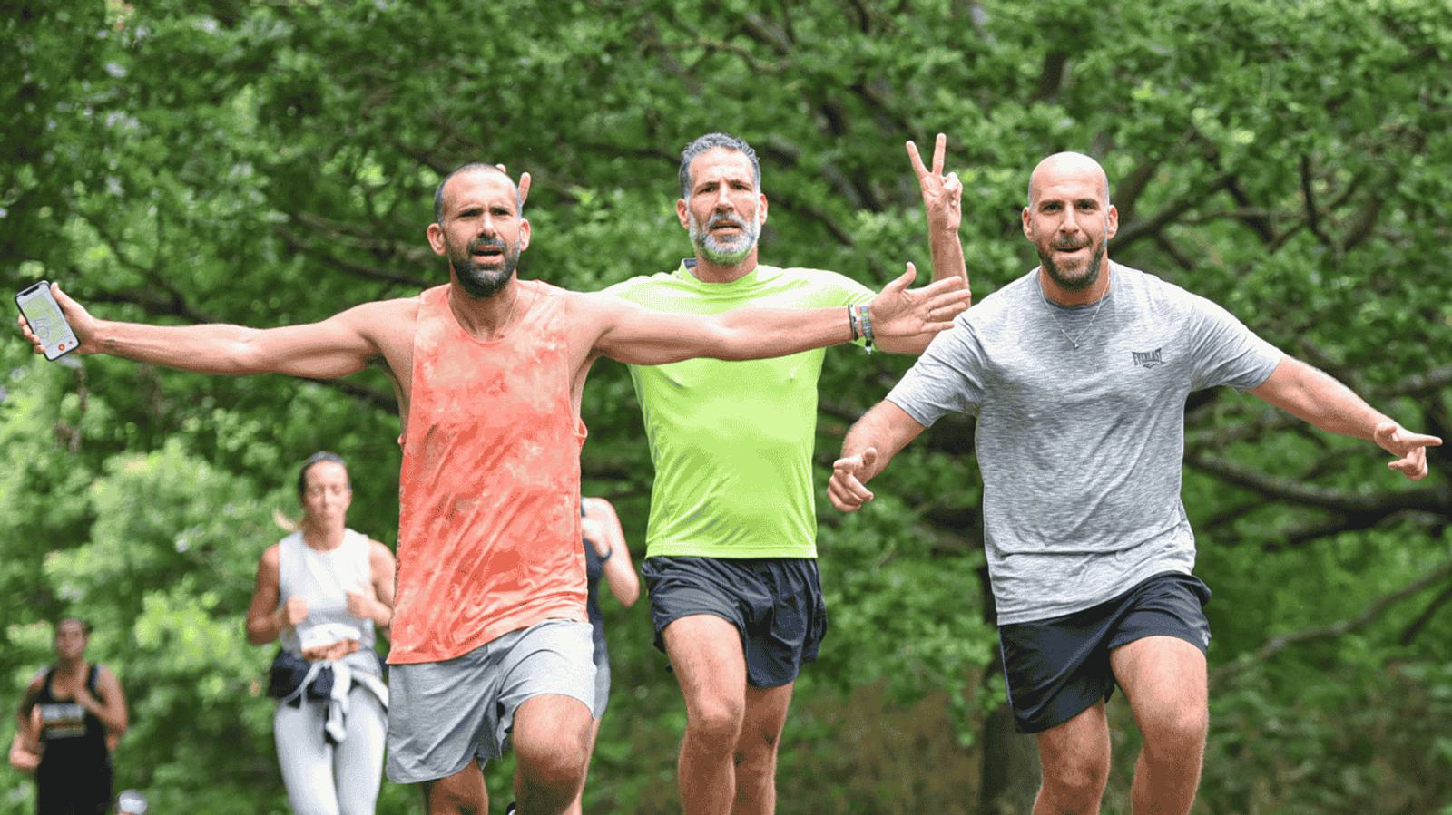 Three men jog together on a tree-lined path, smiling and gesturing energetically. The man on the left wears an orange tank top, the middle man wears bright green, and the right one wears gray. A few other joggers can be seen in the background.