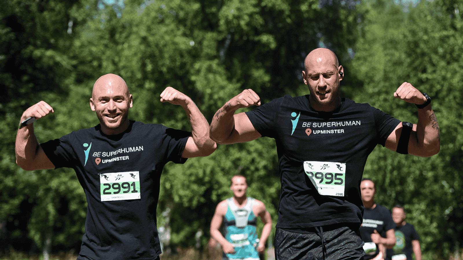Two men with shaved heads smile and flex their arm muscles while running in a race, wearing "Be Superhuman Upminster" T-shirts and race numbers 2991 and 2995. They are on a sunny path with trees and other runners in the background.