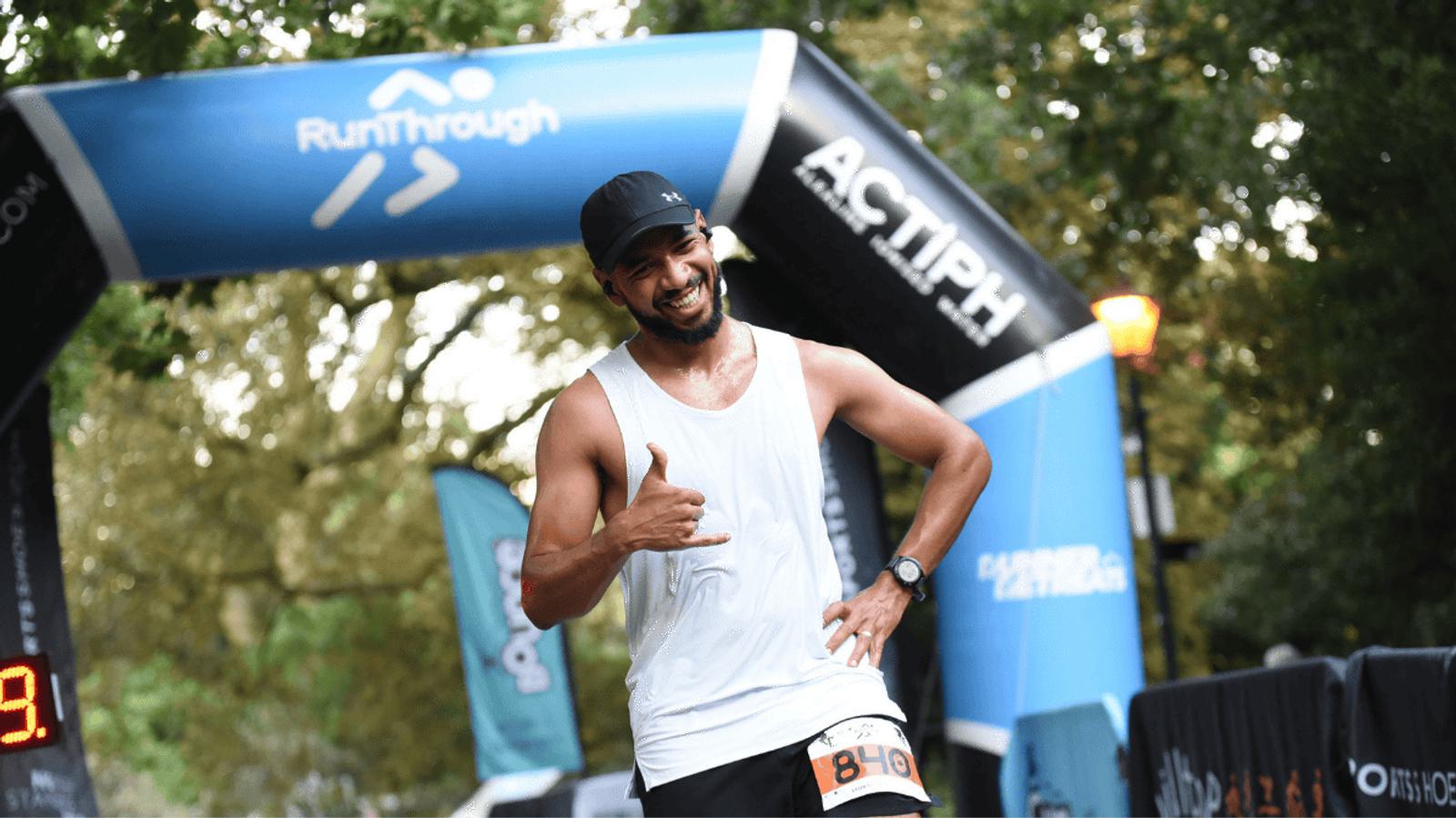 A man in a white tank top and black cap smiles and gestures with his hand as he crosses a race finish line. The finish line banner reads "RunThrough." He wears a race bib numbered 849. Trees and event flags are in the background.