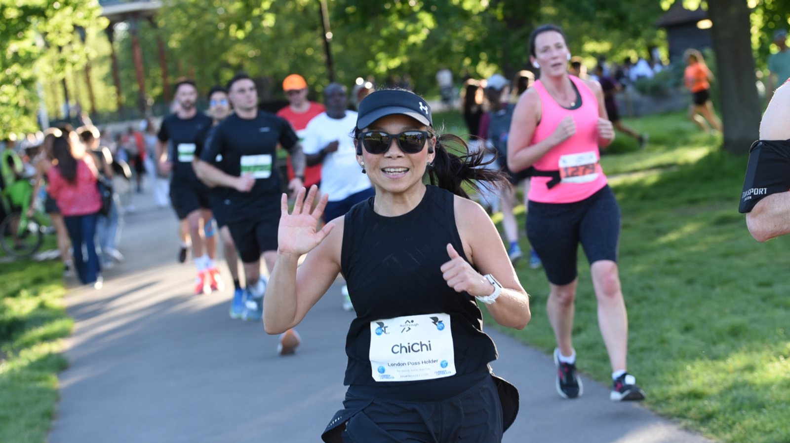 A group of runners competing in a park race. The woman in front, wearing black, is smiling and waving at the camera. Spectators and fellow runners are visible in the background. The sun is shining, and it's likely a warm day.