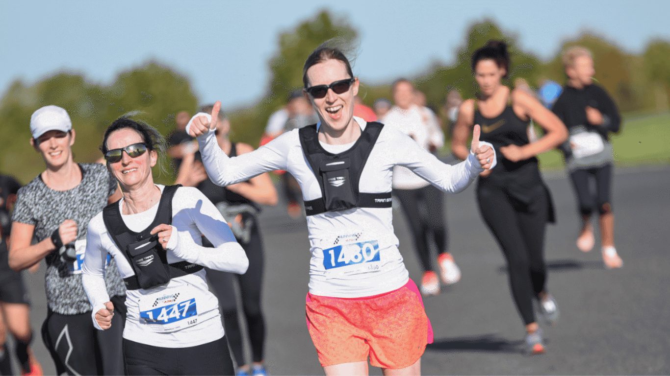 A group of runners, mostly women, participate in an outdoor race on a sunny day. The leading woman in pink shorts is smiling and giving thumbs up. All runners wear numbered bibs. Trees are visible in the blurred background.