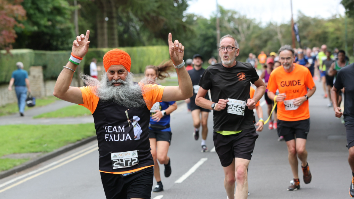 A runner wearing an orange turban and shirt labeled "Team Fauja" energetically gestures while participating in a race. Other runners, including a guide, are visible in the background on a tree-lined street.