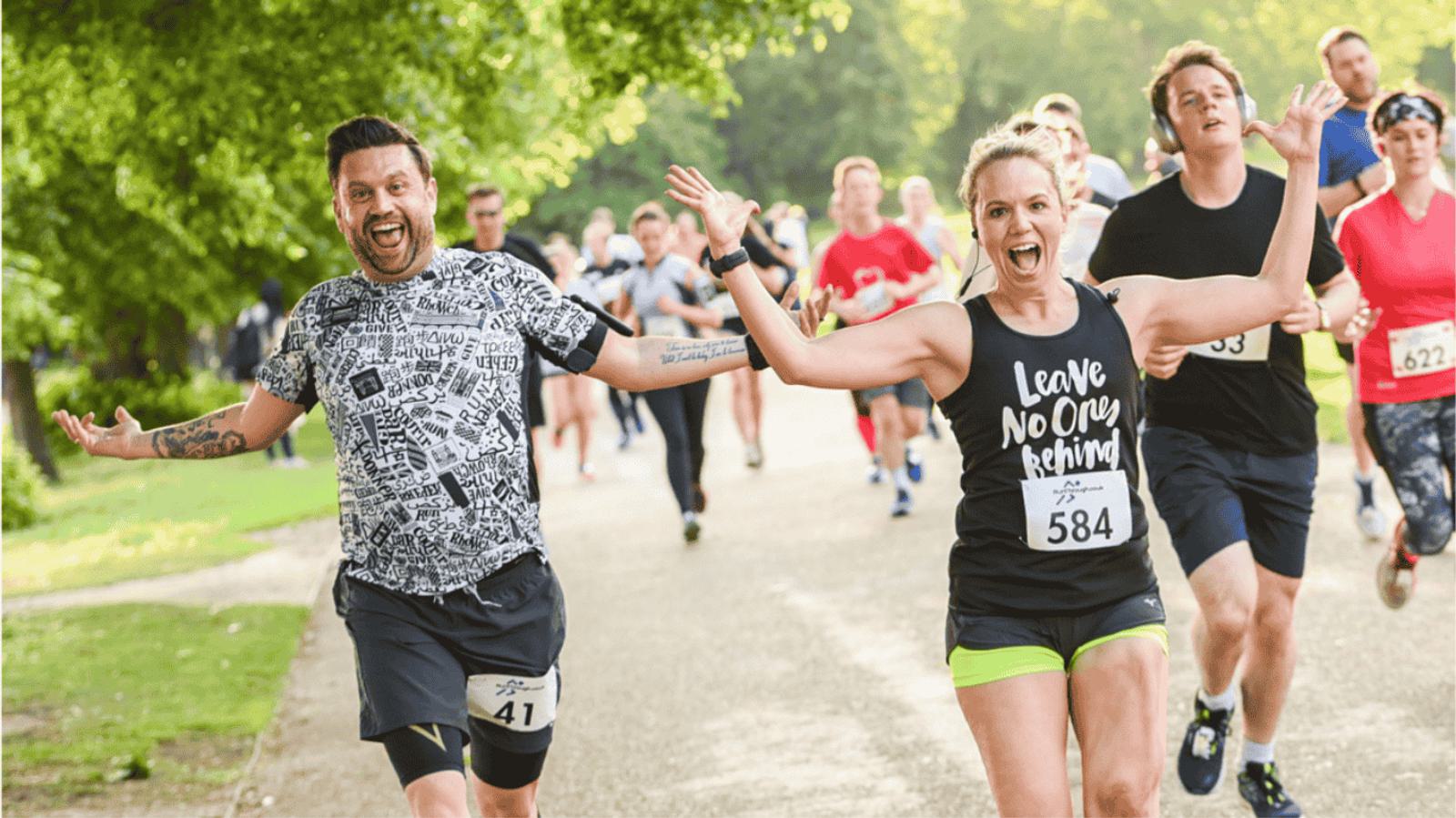 A man and woman enthusiastically run a race, both smiling with arms outstretched. The woman wears a shirt that reads "Leave No One Behind." Other runners follow on a tree-lined path under a sunny sky.