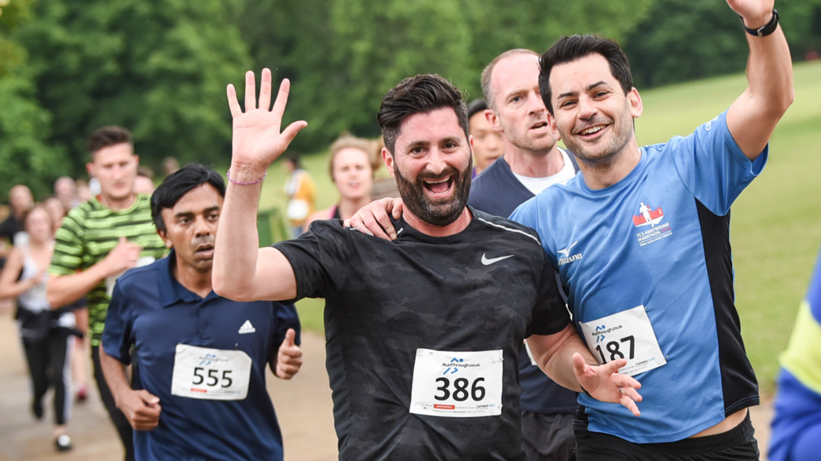 A group of runners with race numbers jog through a park. A person in a black shirt with number 386 waves and smiles, while another in a blue shirt with number 187 runs beside him, also smiling. The background features green trees and grass.