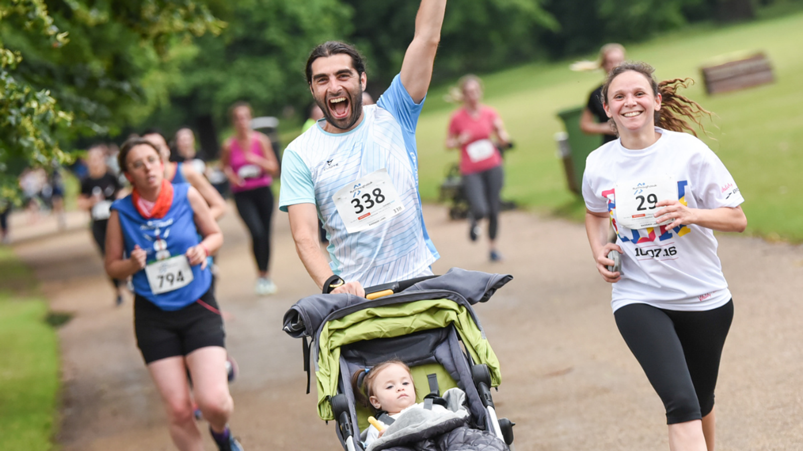 A man joyfully runs while pushing a baby stroller, with a baby inside, during a race. A woman runs beside him, smiling. Other participants are behind them on a tree-lined path. They're wearing numbered race bibs.